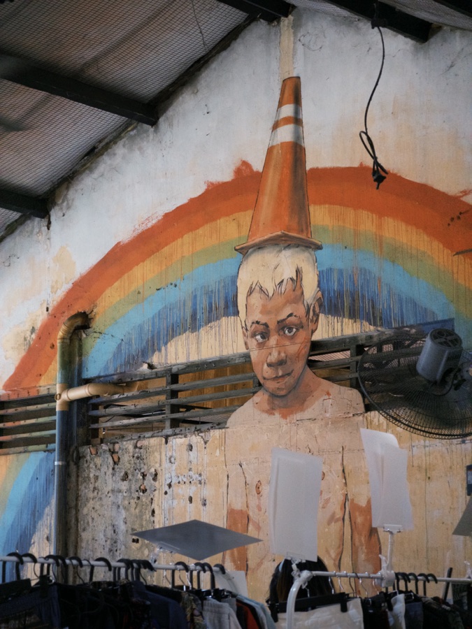 A mural of a person beneath a painted rainbow above clothing racks on a worn wall.