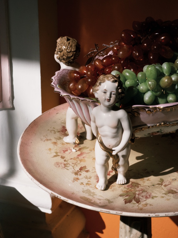 Porcelain figures and decorative grapes on a floral plate lit by sunlight from a window.