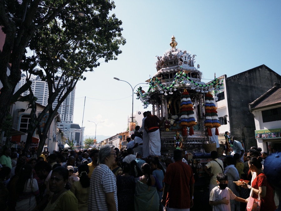 A crowd surrounding a decorated silver carriage with garlands on a tree-lined street.