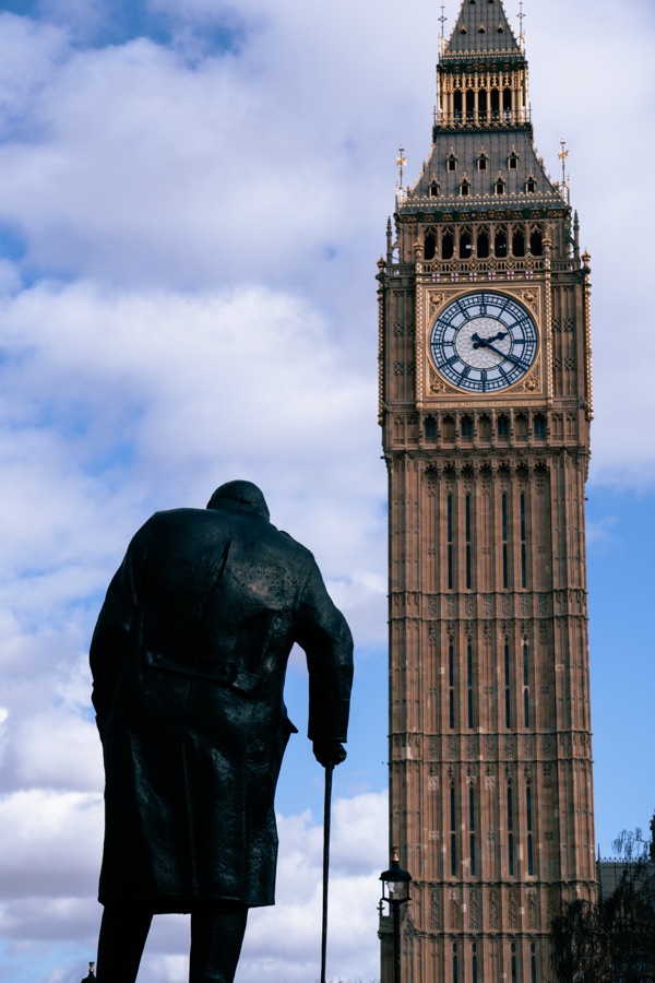 A dark statue in the foreground of an ornate clock tower beneath a cloudy sky.